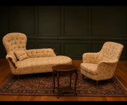 A vintage upholstered chaise lounge and matching armchair in a beige floral pattern, arranged on a patterned area rug with a small wooden side table in front, set against a dark paneled wall.