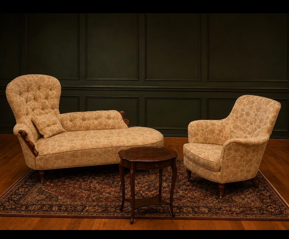 A vintage upholstered chaise lounge and matching armchair in a beige floral pattern, arranged on a patterned area rug with a small wooden side table in front, set against a dark paneled wall.
